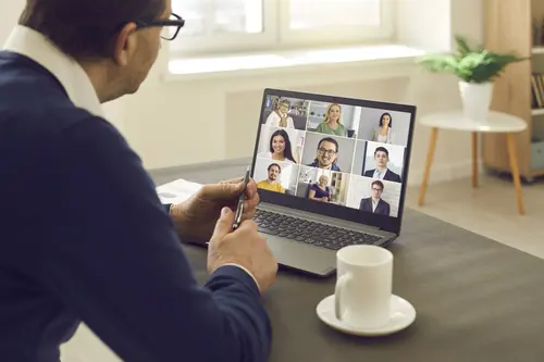 Mann mit Brille sitzt vor Laptop und hält Videokonferenz. Am Bildschirm verschiedene Gesichter