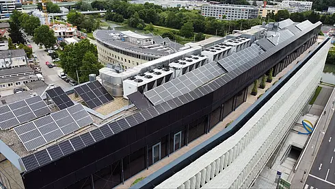 Luftaufnahme eines Gebäudedachs mit umfangreicher Solaranlage, umgeben von städtischer Bebauung.