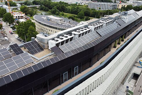 Luftaufnahme eines Gebäudedachs mit umfangreicher Solaranlage, umgeben von städtischer Bebauung.