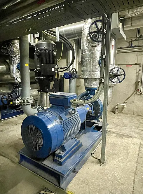 Ein Maschinenraum mit Industrieausrüstung, darunter ein großer blauer Elektromotor, der mit einer Pumpe verbunden ist. Die Anlage umfasst verschiedene Rohre, Ventile und Verbindungsstücke.