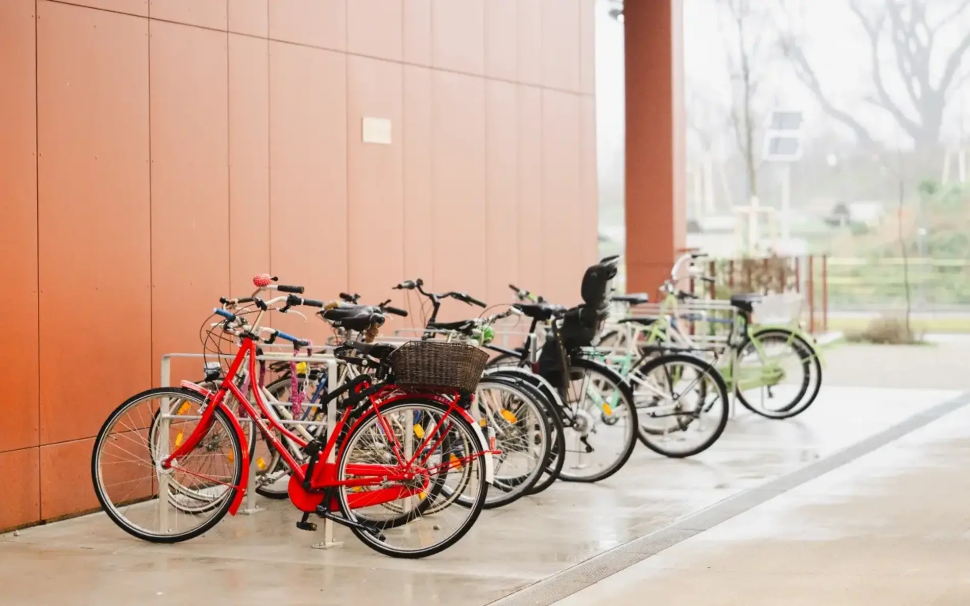 hohe Fahrradständer und verschiedene Fahrräder, die daran hängen. Eins der circa 10 Räder hat einen Kindersitz drauf. Der Radständer ist entlang einer orangenen Gebäudewand aufgestellt.