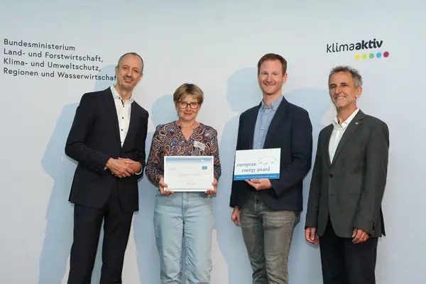 Menschen mit einer Urkunde in der Hand vor einer Fotowand. Sie lächeln in die Kamera.
