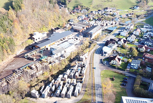 Luftbild eines Holzlagers mit Holzstapeln, Gebäuden, Waldgebiet und Straßen sowie Häusern eines nahegelegenen Dorfes.