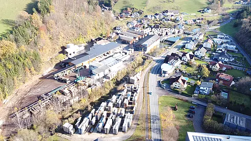Luftbild eines Holzlagers mit Holzstapeln, Gebäuden, Waldgebiet und Straßen sowie Häusern eines nahegelegenen Dorfes.