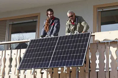 Zwei Männer installieren ein Balkonkraftwerk. Das Solarpanel ist in unsere Richtung ausgerichtet. Der Balkon ist aus Holz.