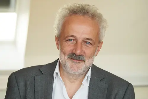 Franz Angerer steht in einem hellen Innenraum und trägt ein strukturiertes Sakko über einem weißen Hemd. Im Hintergrund sind helle Wandflächen, ein Fenster und ein Heizkörper zu sehen. Die Aufnahme zeigt den Oberkörper der Person in stehender Position.