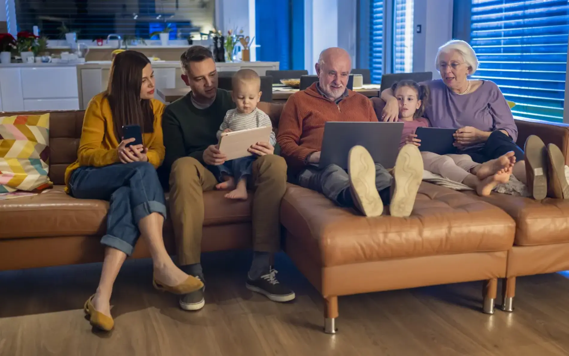 Zwei Eltern mit einem kleinen Kind und zwei Großeltern sitzen auf der Couch. Sie haben alle entweder einen Laptop, ein tablet oder ein Smartphone in der Hand.