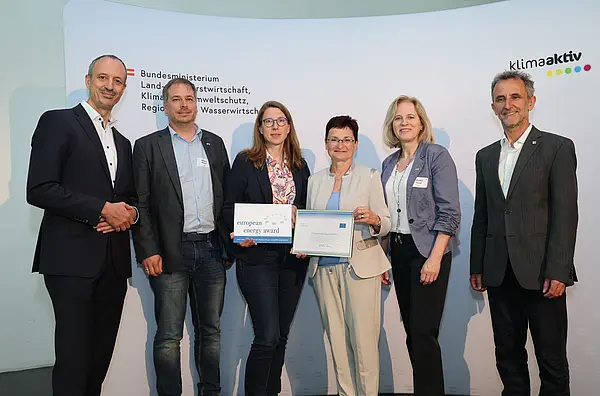Menschen mit einer Urkunde in der Hand vor einer Fotowand. Sie lächeln in die Kamera.