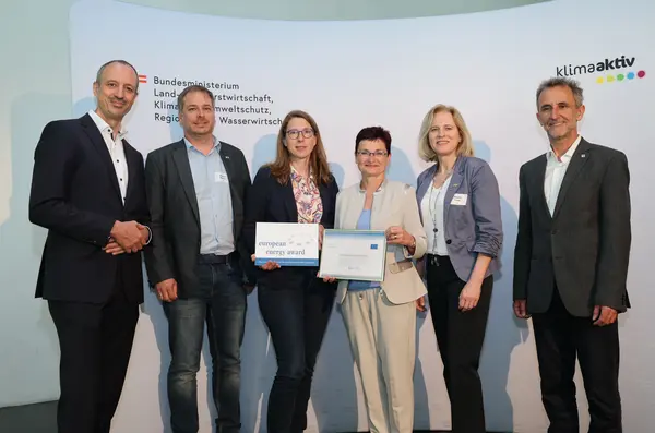 Menschen mit einer Urkunde in der Hand vor einer Fotowand. Sie lächeln in die Kamera.