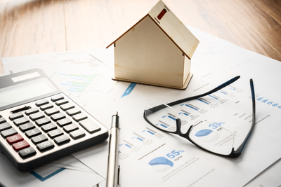 Auf einem großen Papierbogen mit Statistiken liegt ein Taschenrechner, ein Kugelschreiber, eine Lesebrille sowie ein fensterloses Miniaturhaus aus Holz