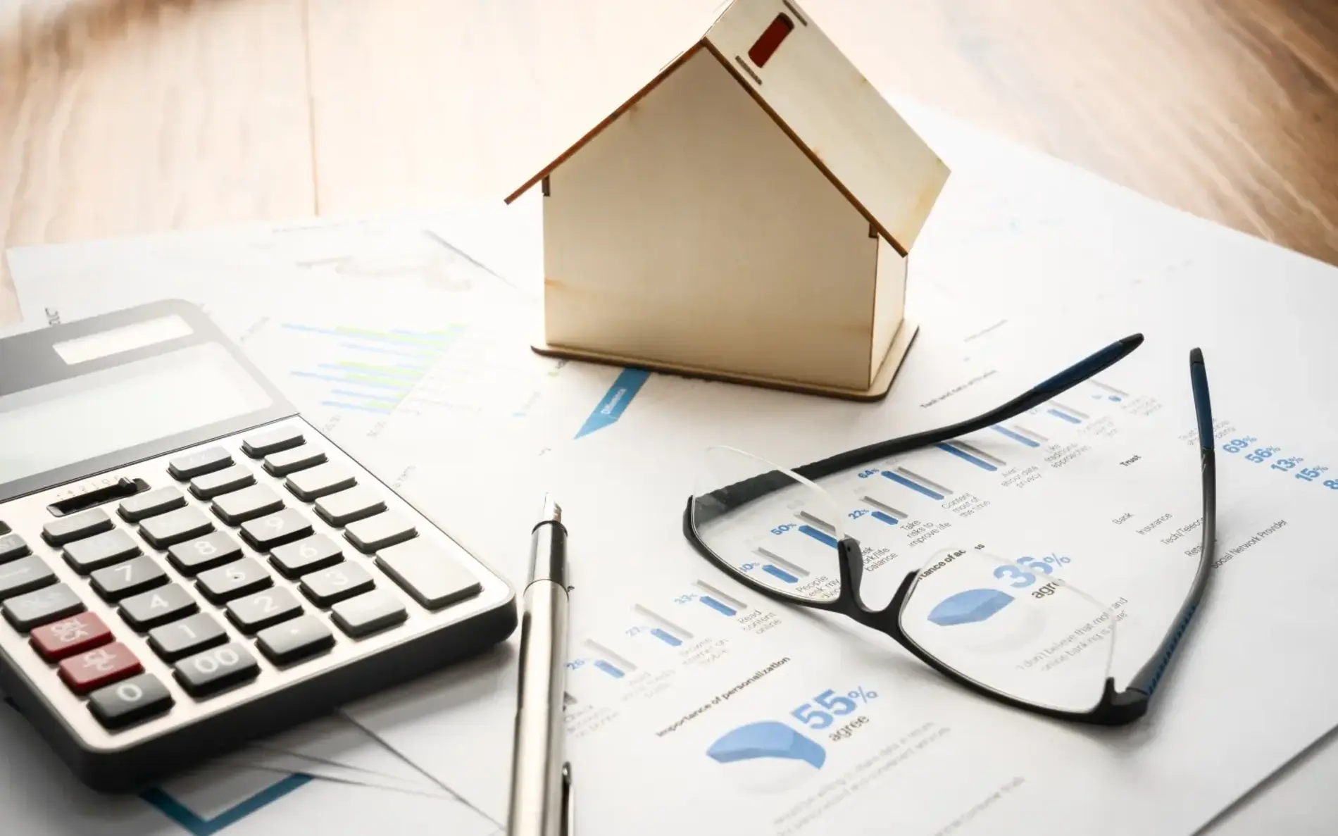 Auf einem großen Papierbogen mit Statistiken liegt ein Taschenrechner, ein Kugelschreiber, eine Lesebrille sowie ein fensterloses Miniaturhaus aus Holz