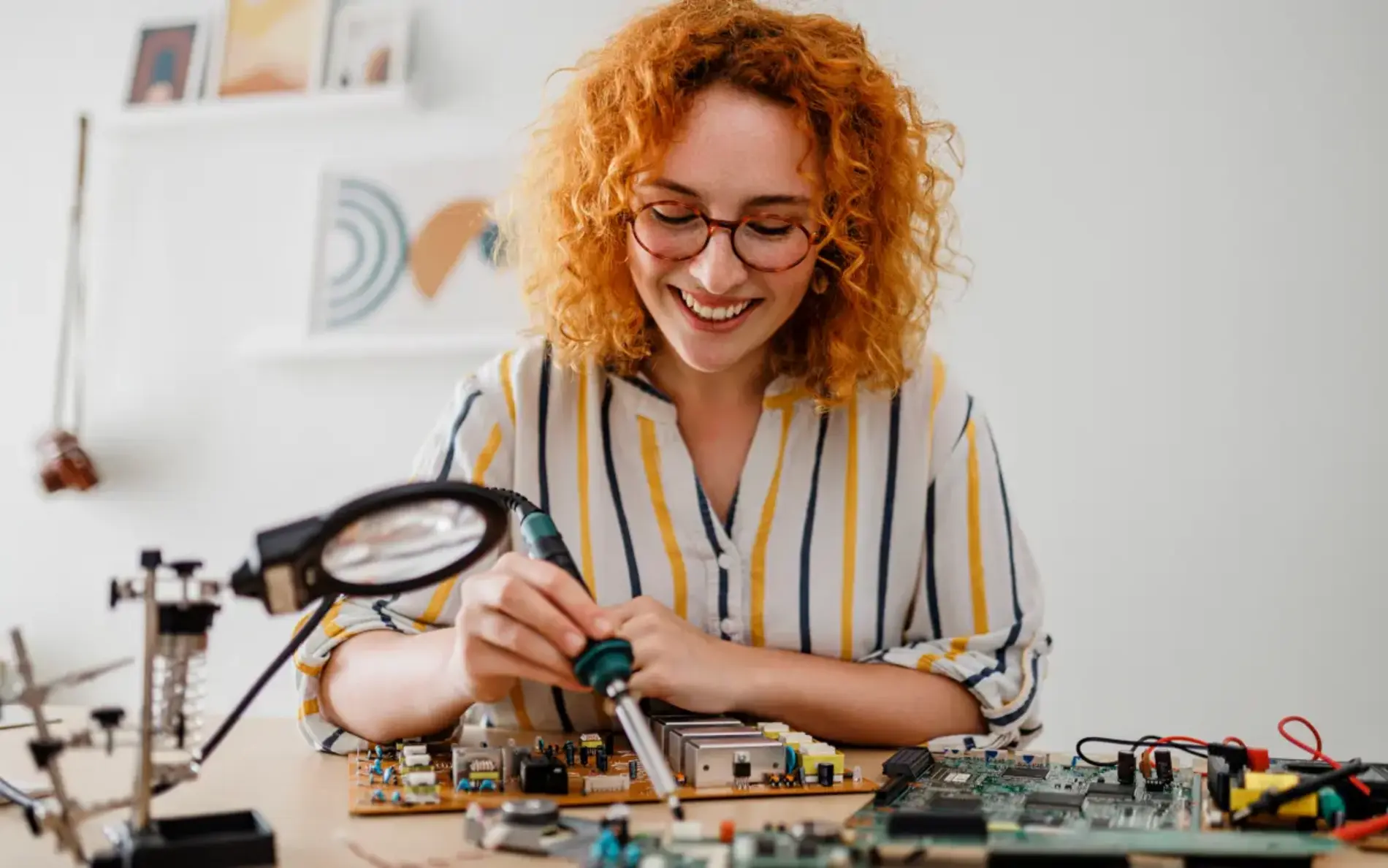 Zu sehen ist eine Frau mit rotem gelockten Haar, die auf einem Schreibtisch sitzt und Elektronikteile zusammenbaut.