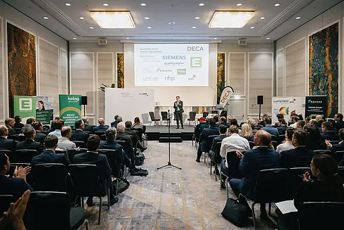 Konferenzszene in einem großen, hellen Raum mit einem Redner auf der Bühne vor einem aufmerksamen Publikum. Im Hintergrund sind ein großer Bildschirm mit Firmenlogos wie DECA, SIEMENS, Energie Steiermark und RHP Technology GmbH sowie mehrere Banner und Poster zu sehen, die verschiedene Unternehmen und Organisationen präsentieren.