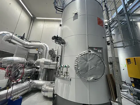 Ein industrieller Bereich mit großen zylindrischen Tanks und umfangreicher Rohrleitung. Die Rohre sind isoliert und mit verschiedenen Maschinen verbunden. Rechts im Bild führt eine Leiter zu einer Plattform. Die Umgebung wirkt sauber und gut organisiert, typisch für eine Anlage zur Verarbeitung von Flüssigkeiten oder Gasen unter Druck.