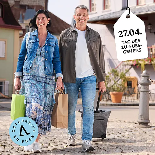 Ein Paar geht mit Rollkoffer und Papiersackerl durch einen kleinstädtischen Ortskern. Rechts im Bild ein reinhängendes Schild mit "27.04. Tag des Zu-Fuß-Gehens"