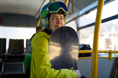 Frau mit Snowboard, dicker Winterjacke in Signalgelb und Skihelm steht in einem öffentlichen Bus.