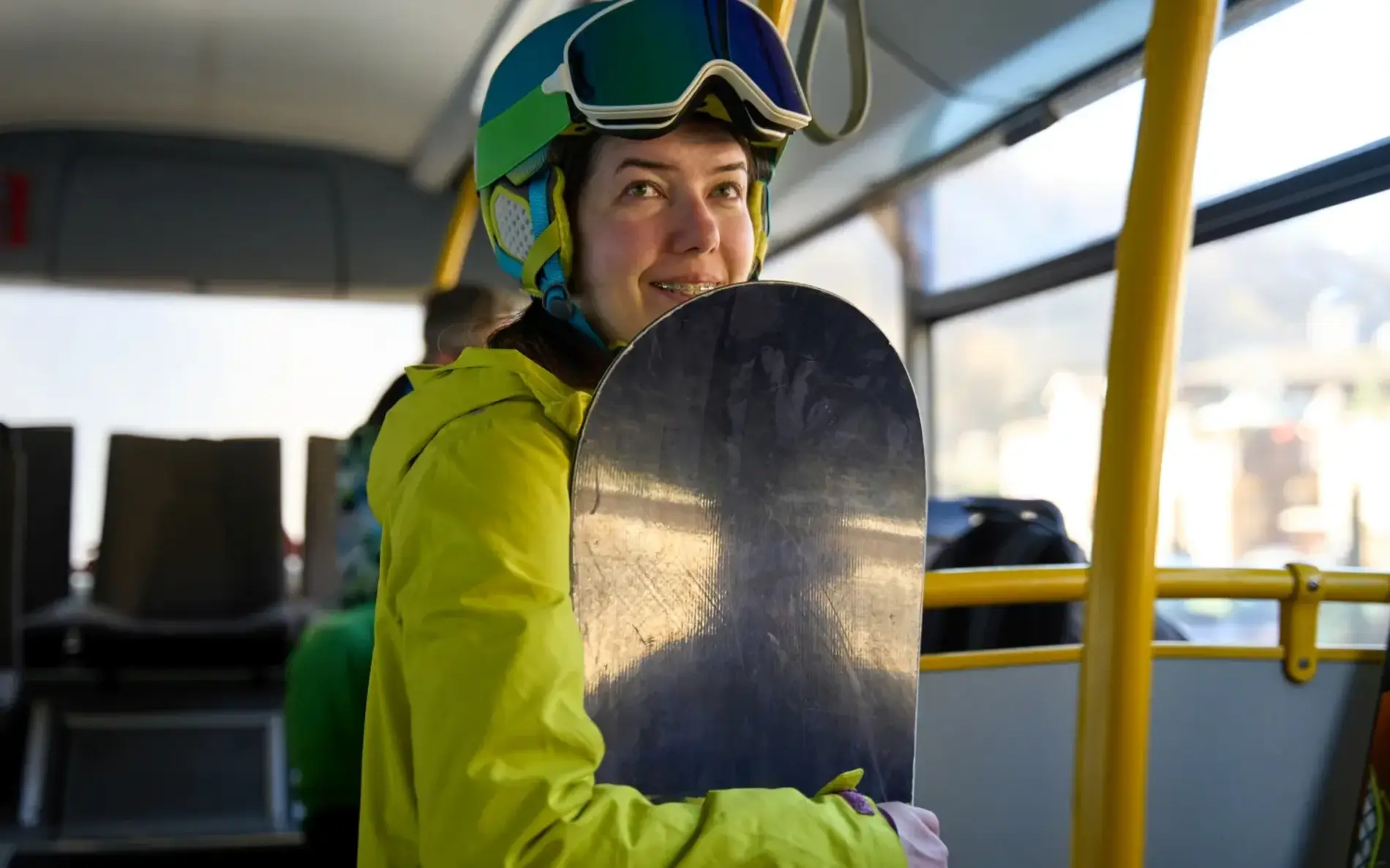 Frau mit Snowboard, dicker Winterjacke in Signalgelb und Skihelm steht in einem öffentlichen Bus.