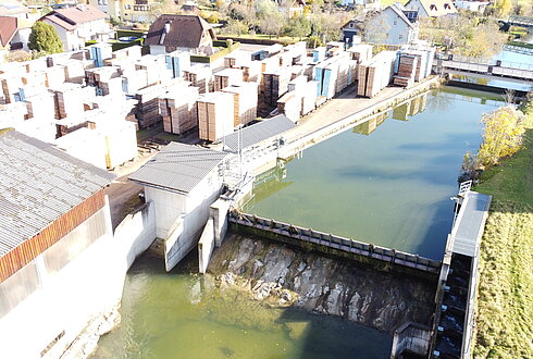 Ein Wasserlauf verläuft neben einem Bereich mit gestapeltem Holz und angrenzenden Gebäuden in einem Wohngebiet.
