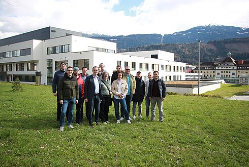 Mehrere Personen stehen auf einer Wiese vor einem mehrstöckigen Gebäude mit großen Fenstern. Im Hintergrund sind weitere Gebäude und schneebedeckte Berge unter teils bewölktem Himmel zu sehen. Gesichter sind unkenntlich gemacht