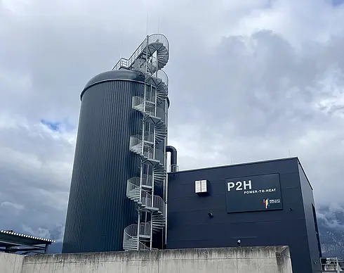 Industrieanlage mit einer großen zylindrischen Struktur auf der linken Seite und einem rechteckigen Gebäude auf der rechten Seite. Die Zylinderstruktur ist mit einer Wendeltreppe umgeben, die nach oben führt. Auf dem Gebäude befindet sich ein Schild mit der Aufschrift „P2H POWER-TO-HEAT“ sowie weiteren kleineren Texten. Im Hintergrund ist ein bewölkter Himmel zu sehen.