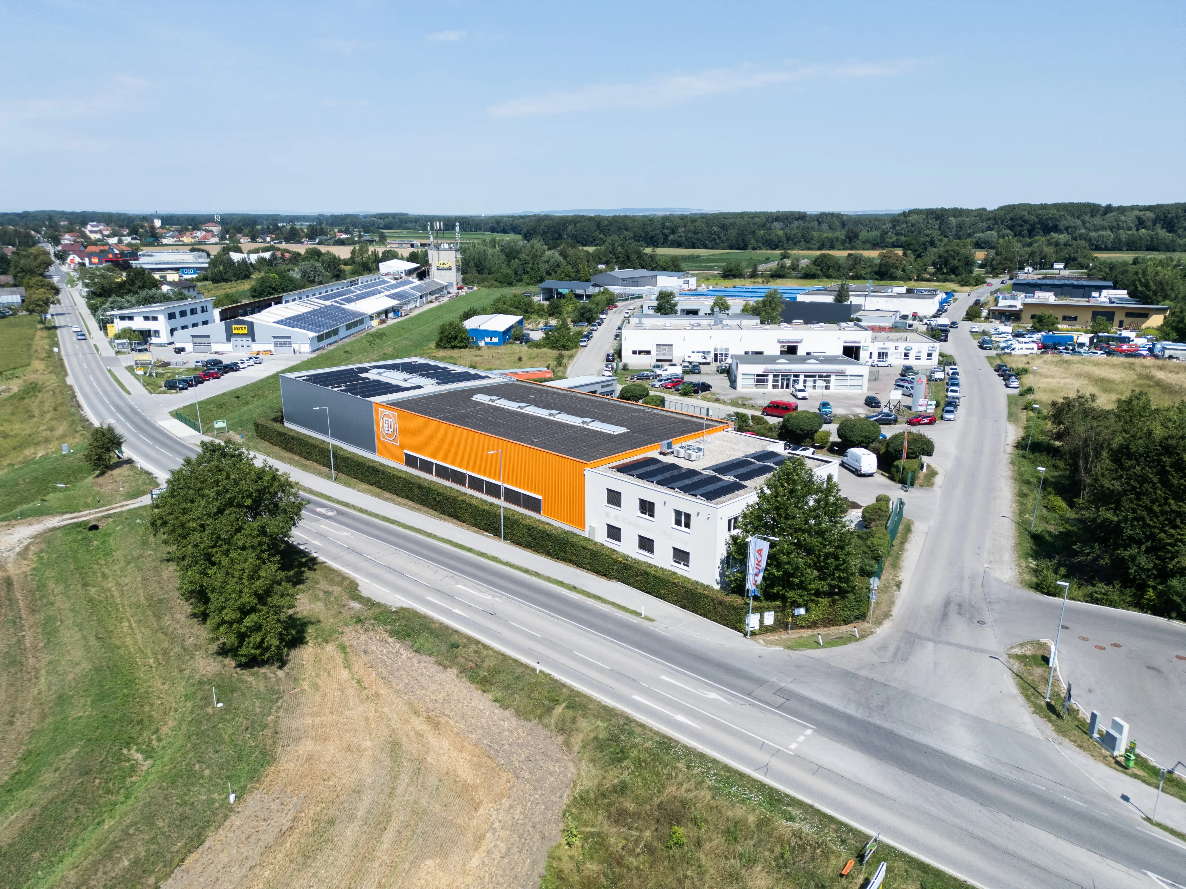 Luftaufnahme eines modernen Firmengebäudes mit orangefarbener Fassade und Photovoltaikanlage auf dem Dach, umgeben von Straßen und weiteren Gewerbebauten in einer grünen Landschaft.