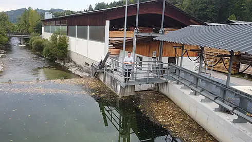 Wasserlauf neben einem Sägewerksgebäude, eine Person steht auf einer Plattform am Ufer.
