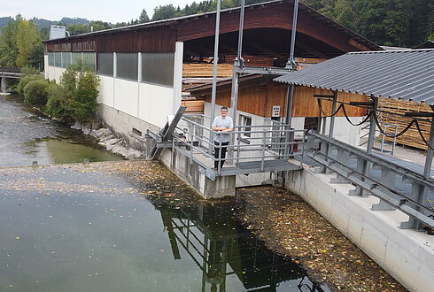 Wasserlauf neben einem Sägewerksgebäude, eine Person steht auf einer Plattform am Ufer.