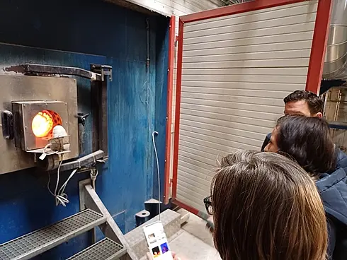 Gruppe von Personen in einem industriellen Umfeld beobachtet einen geöffneten Ofen mit orangefarbenem Glühen im Inneren.