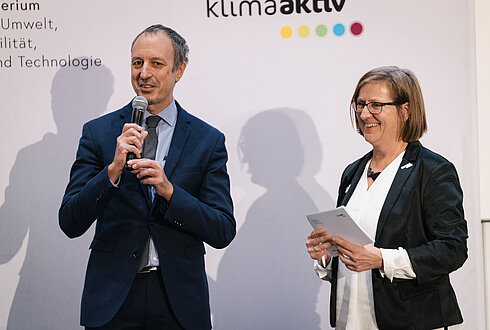 Zwei Personen vor einem Hintergrund mit den Logos ‚klimaaktiv‘ und ‚Bundesministerium für Umwelt, Mobilität, Innovation und Technologie‘, eine Person mit Mikrofon, die andere mit Papier