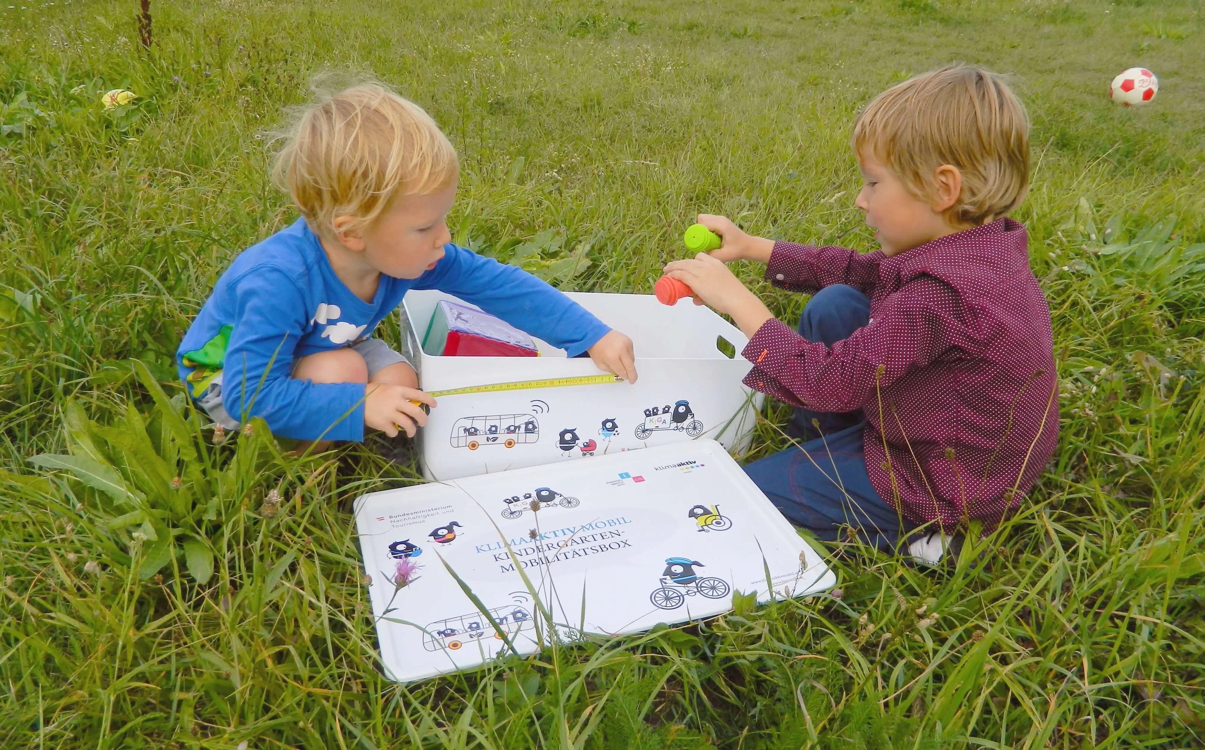 Zwei Kinder spielen öffnen die Kindergarten-Mobilitätsbox. Sie sitzen dabei auf der Wiese.