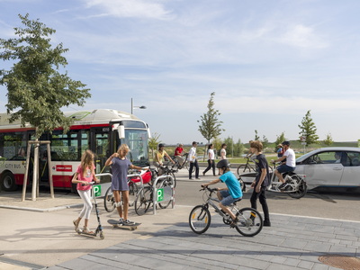 Ein breiter Geh- und Radweg mit Kindern auf Roller und Rad. Dahinter eine Straße mit Bus und E-Auto