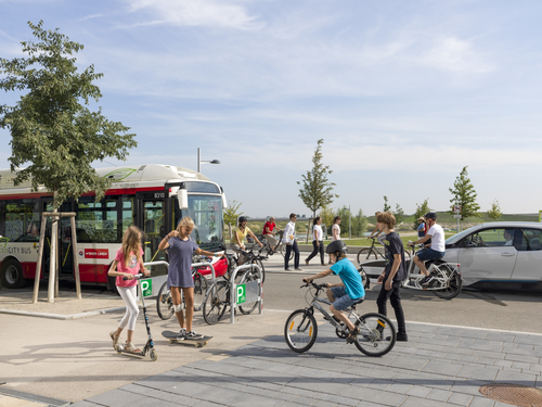 Ein breiter Geh- und Radweg mit Kindern auf Roller und Rad. Dahinter eine Straße mit Bus und E-Auto