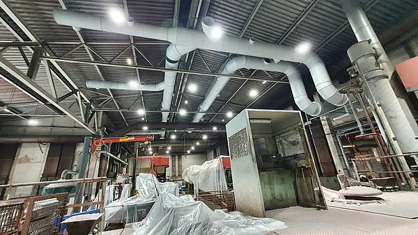 Industrielle Lüftungsrohre, die an der Decke einer großen Halle installiert sind, mit Maschinen und abgedeckten Materialien im Vordergrund.