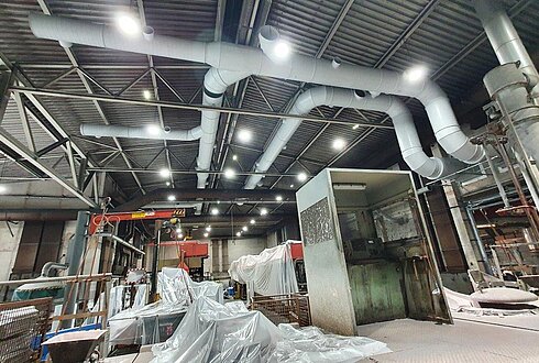 Industrielle Lüftungsrohre, die an der Decke einer großen Halle installiert sind, mit Maschinen und abgedeckten Materialien im Vordergrund.