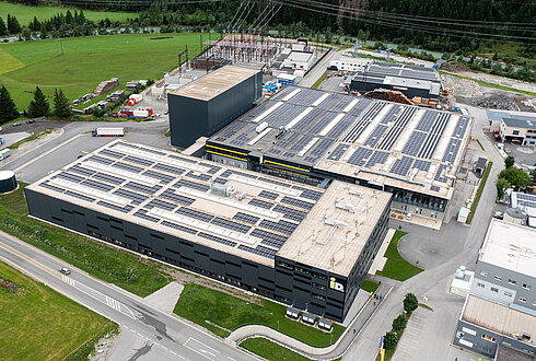 Luftaufnahme eines großen Industriekomplexes in einem Tal, umgeben von schneebedeckten Bergen. Mehrere Gebäude und Straßen sind sichtbar, sowie grüne Felder im Vordergrund.