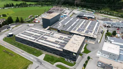 Luftaufnahme eines großen Industriekomplexes in einem Tal, umgeben von schneebedeckten Bergen. Mehrere Gebäude und Straßen sind sichtbar, sowie grüne Felder im Vordergrund.