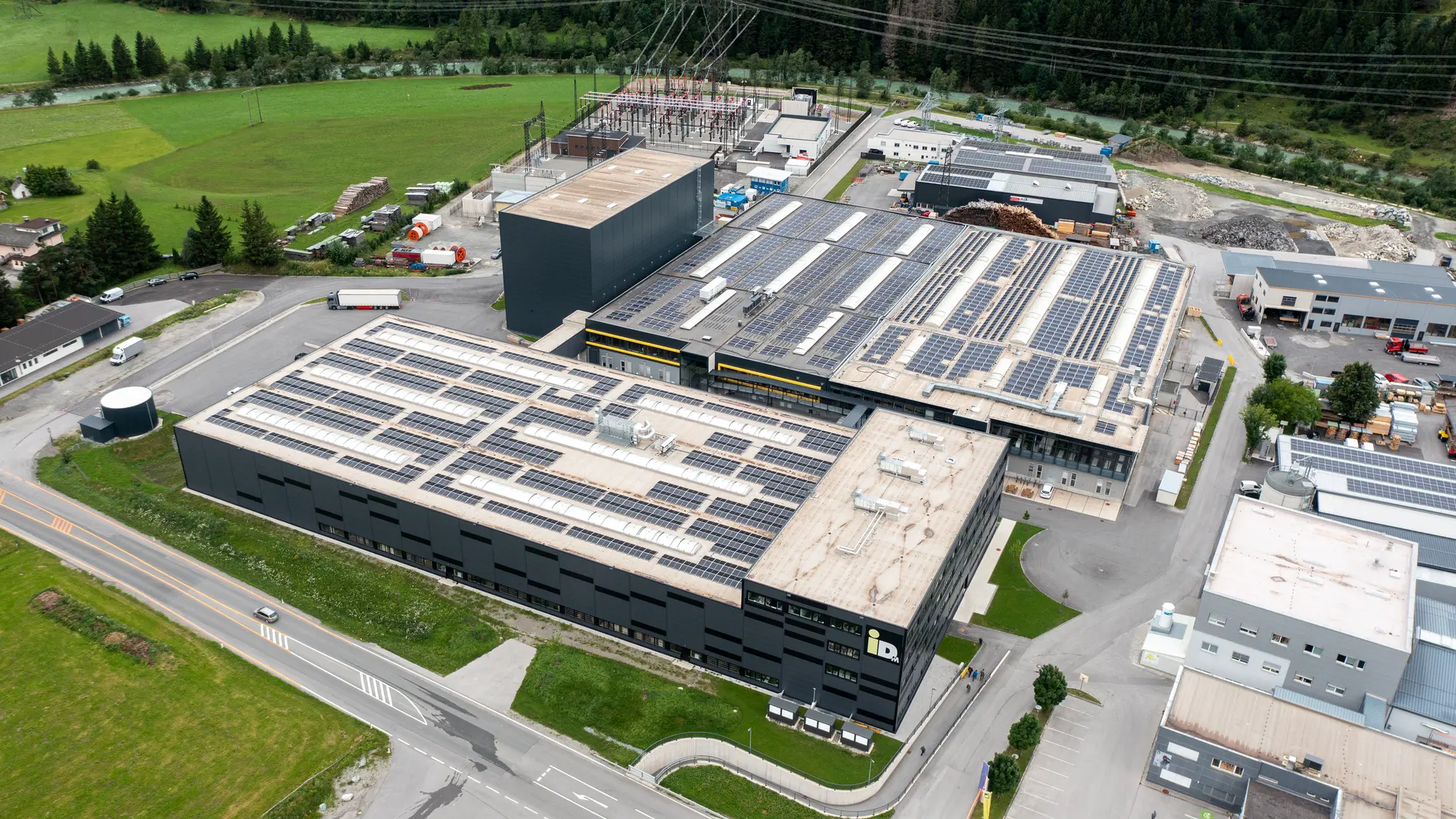 Luftaufnahme eines großen Industriekomplexes in einem Tal, umgeben von schneebedeckten Bergen. Mehrere Gebäude und Straßen sind sichtbar, sowie grüne Felder im Vordergrund.