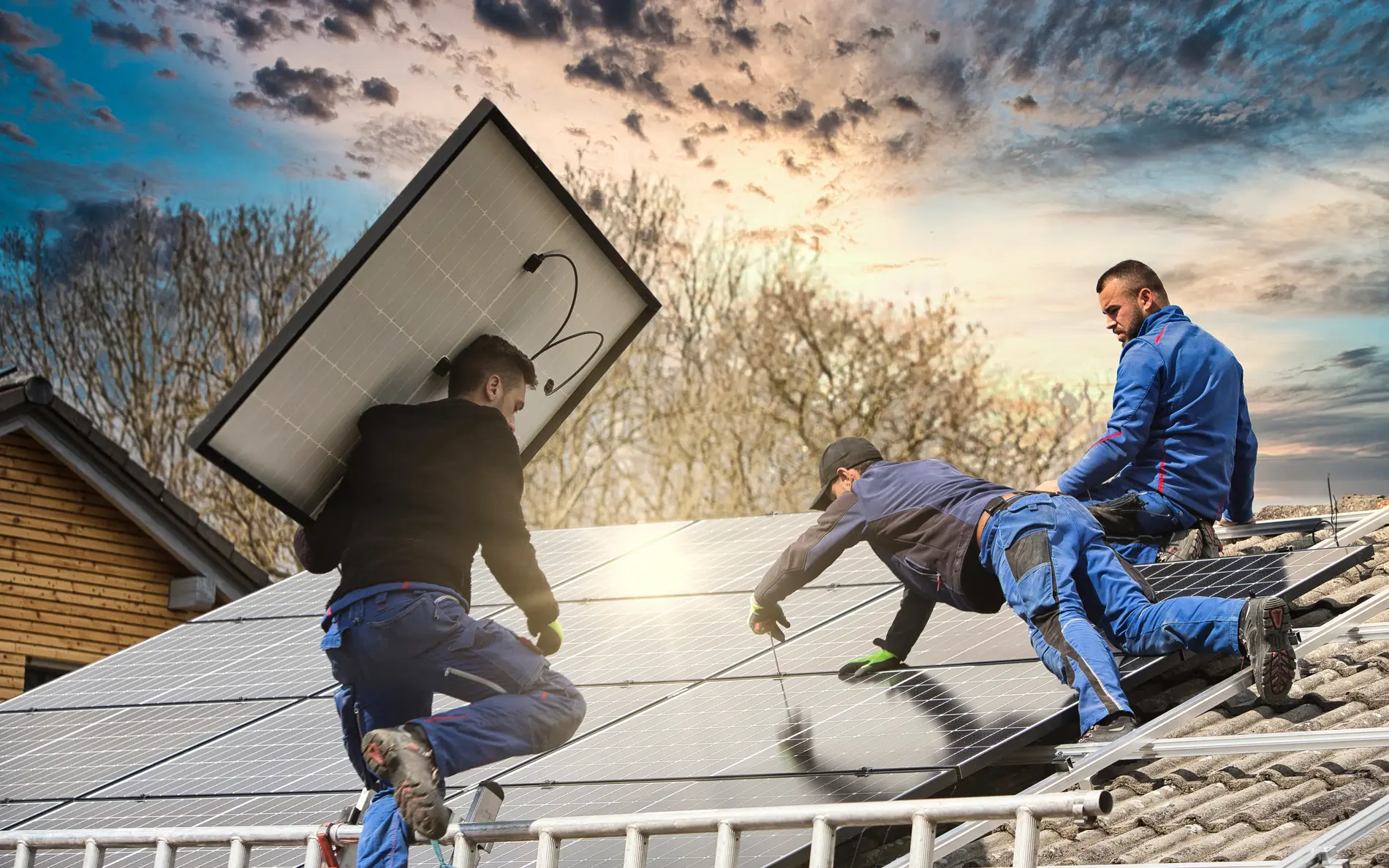 Drei Männer befestigen Solarpanele am Dach. Die Hälfte der Photovoltaikanlage ist bereits fertig. Einer der Männer klettert gerade mit einem Panel auf der Schulter die Leiter hoch.