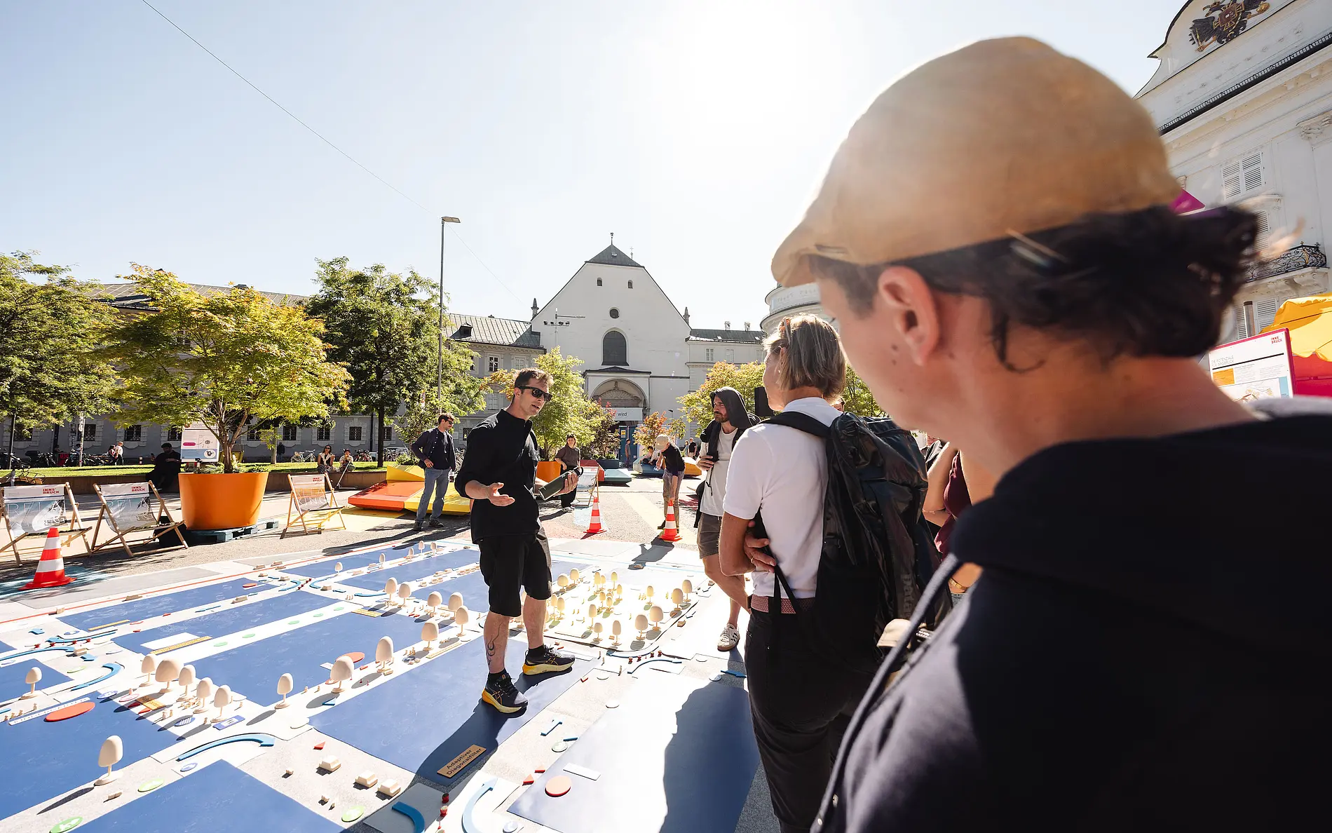 Personen schauen auf eine Bodeninstallation, die einen verkehrsberuhigten Straßenblock darstelllt. Sie befinden sich im Freien, im Hintergrund Gebäude und Pflanzen
