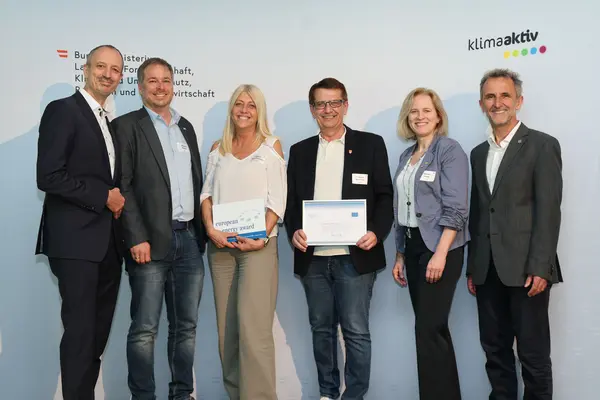 Menschen mit einer Urkunde in der Hand vor einer Fotowand. Sie lächeln in die Kamera.