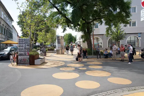 Straßenbemalung mit großen gelben Punkten, im Hintergrund einzelne Menschen, die sich auf Bänken unter Bäumen ausruhen.