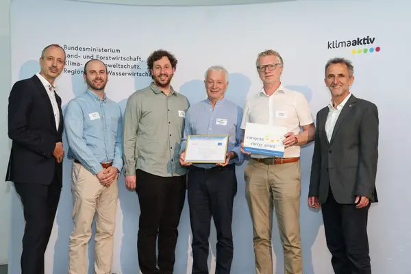 Menschen mit einer Urkunde in der Hand vor einer Fotowand. Sie lächeln in die Kamera.