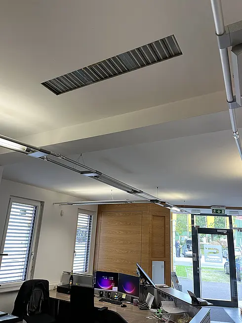 Innenansicht eines modernen Büros mit Schreibtischen, Monitoren und großen Fenstern. Lüftungsschlitz an der Decke