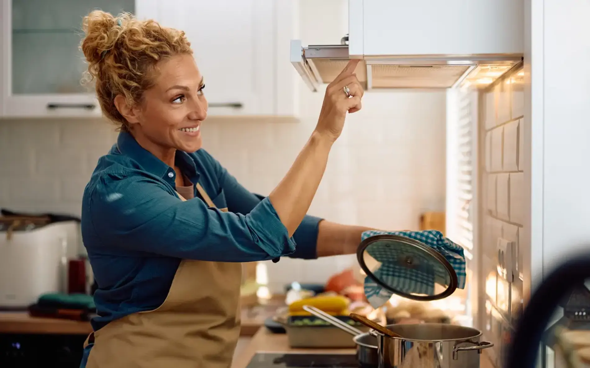 Eine Frau mit gelocktem blonden Haaren und einer Kochschürze schaltet beim kochen die Dunstabzugshaube ein. 