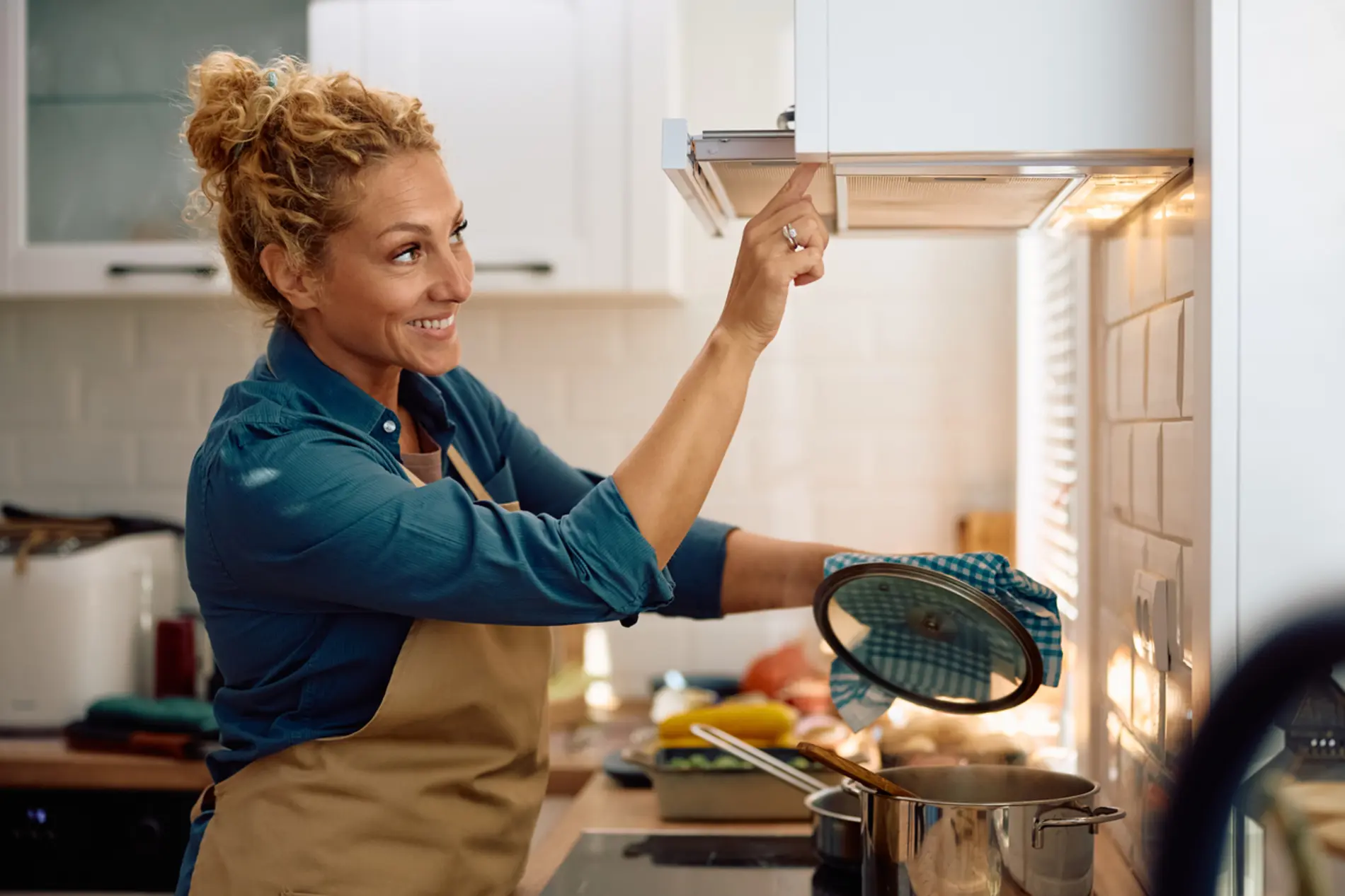 Eine Frau mit gelocktem blonden Haaren und einer Kochschürze schaltet beim kochen die Dunstabzugshaube ein. 