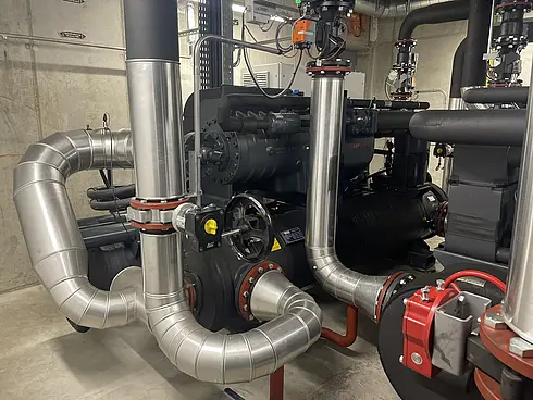 Wärmepumpe, die im Biomasse Heizkraftwerk der Hall AG zum Einsatz kommt. in Maschinenraum mit Industrieausrüstung, darunter große Metallrohre, Ventile und Maschinen. Die Rohre sind isoliert und mit verschiedenen Komponenten verbunden.