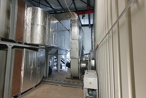 Ein großes Lüftungssystem mit wärmegedämmten Leitungen und Kupfer-Wärmetauschern in einer Industriehalle. Im Vordergrund ist ein elektrisch betriebenes Gerät zu sehen.