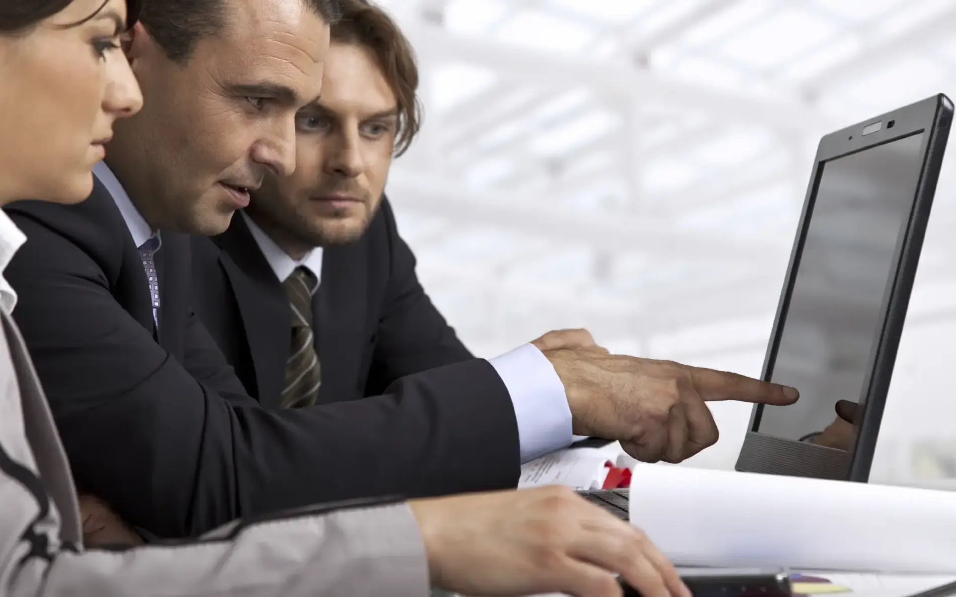 Drei Personen starren auf einen Laptop-Bildschirm, einer im Anzug zeigt mit Finger darauf
