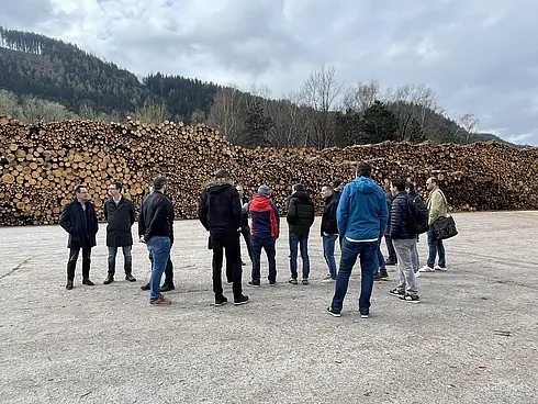 Eine Gruppe von Personen steht im Freien vor einem großen Stapel geschnittener Holzstämme. Die Szene spielt sich vermutlich in einem Holzlager oder an einem Holzernteplatz ab. Im Hintergrund sind bewaldete Hügel unter einem bewölkten Himmel zu sehen.