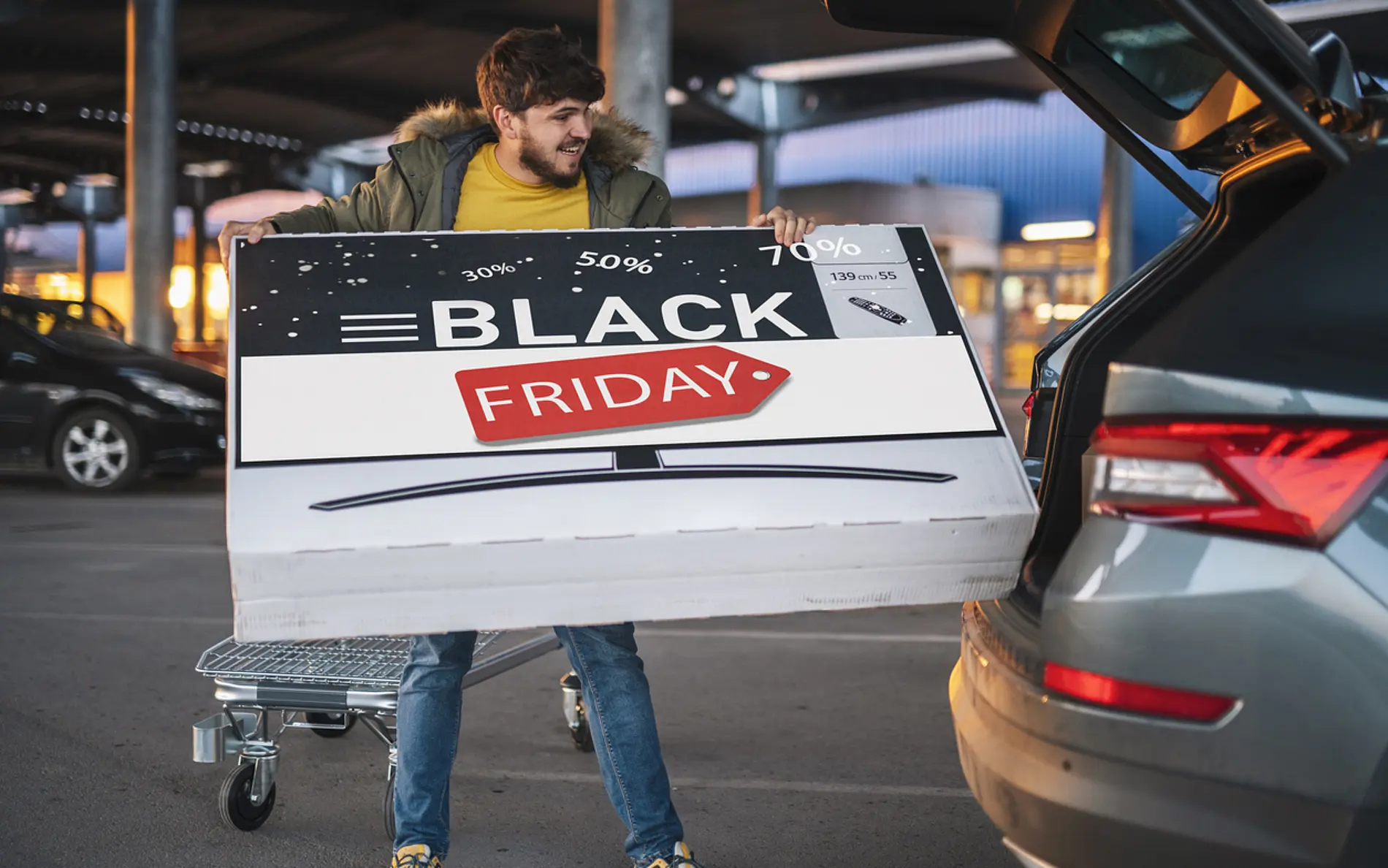 Ein Mann lädt ein großen Karton, bei welchem Ein Fernseher und der Schriftzug "Blackfriday" abgebildet ist, in den Kofferraum
